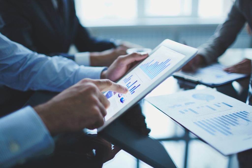 Photo of a person holding a tablet showing bar charts, with a backdrop of a meeting table with papers of charts.